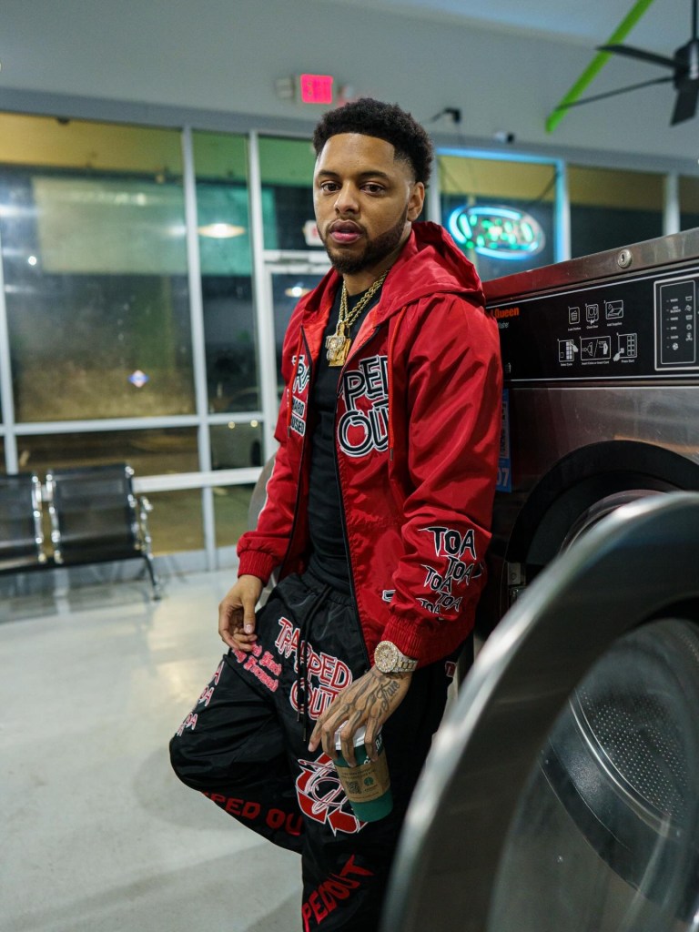 A young man wearing a red jacket and black pants stands by a washing machine in a laundromat, holding a coffee cup.