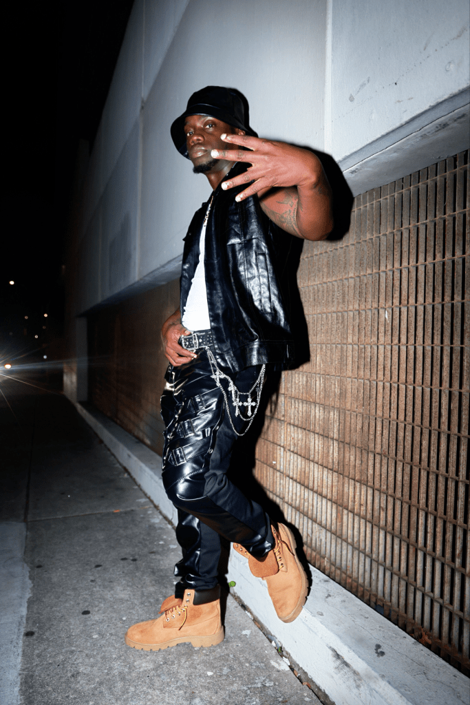 A person stands against a textured wall at night, wearing a black sleeveless jacket, black pants with straps, and tan boots. They are making a hand gesture with four fingers showing.