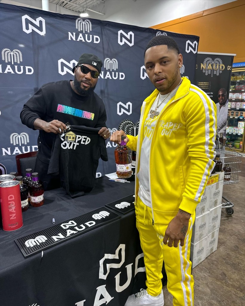 Two men posing at a promotional event for NAUD, one wearing a black shirt and sunglasses holding a shirt, while the other in a yellow tracksuit holds a bottle.