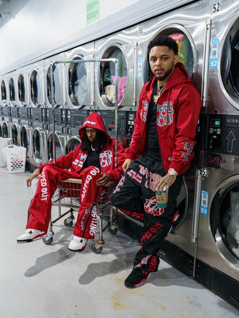 Two individuals in vibrant streetwear outfits pose inside a laundromat, with washing machines in the background. One person sits in a shopping cart while the other leans against a machine, both wearing hoodies and branded apparel.